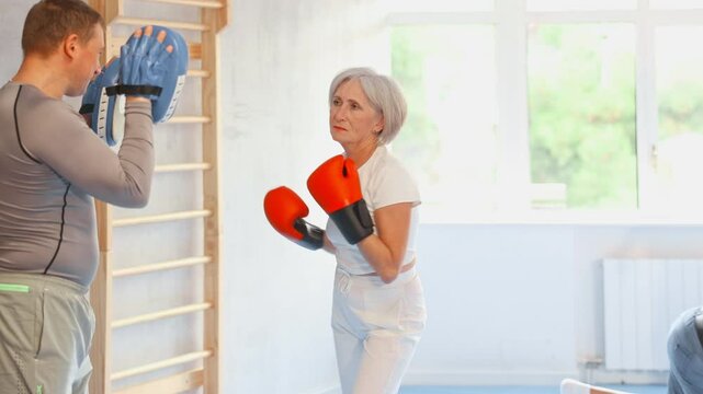 Senior lady and male couch are boxing in gym, man helps student to work out force of blow. Modern positive family chooses active hobby