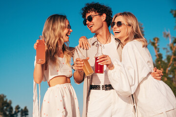 Young stylish friends posing at the seaside. Fashion man and two cute female dressed in casual summer clothes. Smiling models having fun. Cheerful women and guy outdoors, Hold and drink lemonade