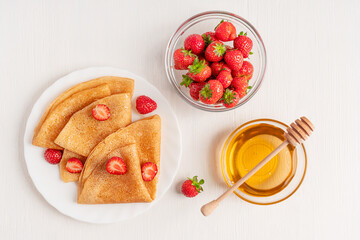 Flat lay of sweet fried french crepe or blini dessert snack decorated with sliced berries served on round plate on white wooden table with bowl of honey with dipper and fresh juicy red strawberries