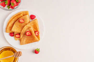 Top view of sweet folded fried crepe or blini dessert snack decorated with sliced strawberries served on round plate on white wooden background with copy space with glass bowl of honey for breakfast