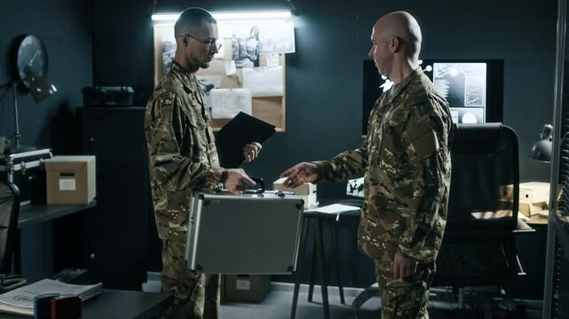 Medium long shot of adult sergeant in camouflage uniform giving suitcase with secret documents to male colleague in command center