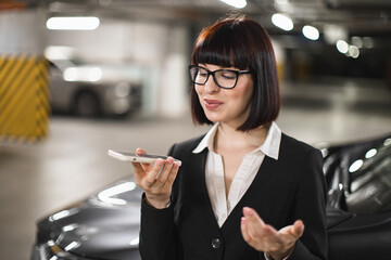 Young adult Caucasian woman recording voice message on smartphone in a modern car park....