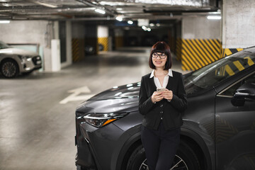 Fototapeta premium Young Caucasian business woman in office attire using smartphone in indoor car park, leaning against car with confident expression, emphasizing communication and modern lifestyle in urban setting.