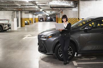 Caucasian female business professional stands in parking garage holding laptop. Young adult focused...