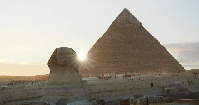Sunset sunburst rays between the silhouettes of the Great Sphinx and Khafre Pyramid in Giza, with crowds of tourists behind, Egypt