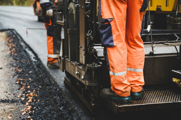 Construction worker standing at the screed of an asphalt paver during the asphalt layering process, ensuring precision and quality in the road paving operation.