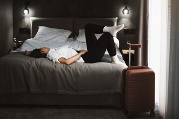 Sad woman sits on the floor, feeling overwhelmed and lonely. Female with suitcase having frustration or after abuse. Dark hotel room with curtains, grief