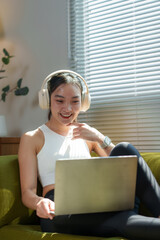 Smiling young asian woman wearing headphones and working on a laptop while sitting on a comfortable...