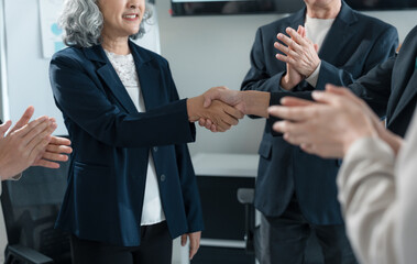 Two businesswomen shaking hands after successfully closing a deal, while colleagues clapping and...