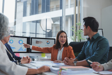 Asian businesswoman leading a meeting with her colleagues, pointing at charts displayed on a computer screen, discussing strategy and analyzing data in a modern office
