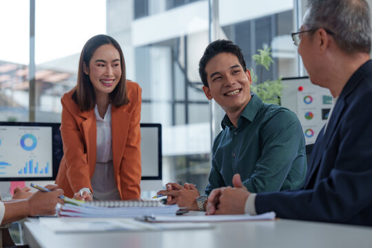 Asian businesspeople collaborating in a modern office, discussing project strategy while analyzing financial charts and graphs on a computer and documents