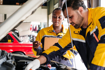 Fototapeta premium Two professional auto mechanics in a repair shop inspecting a car engine. A woman holds a clipboard while a male technician works under the hood. Teamwork and automotive service industry concept.