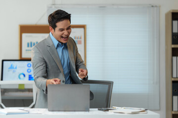 Asian businessman in a gray suit and blue shirt is excitedly celebrating a business achievement while working on his laptop in a modern office, expressing joy and accomplishment
