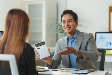Smiling financial advisor discussing investment strategies while showing a calculator to the client during a meeting in a professional office setting, focusing on financial planning and analysis
