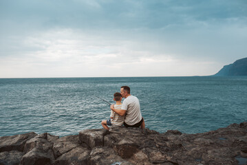 Rear view of father and son fishing in the sea. Sitting on rock holding fish rod.