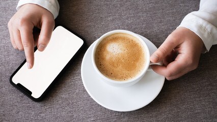 Top-down view of a woman's hands holding a white coffee cup on a saucer and a smartphone mockup with a blank white screen. Cafe restaurant, digital lifestyle, technology, communication, relaxation. 