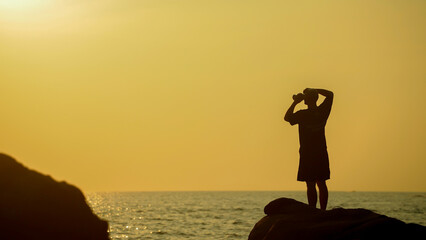 A person stands atop a rock by the ocean, gazing at the vibrant sunset. The warm colors reflect on...