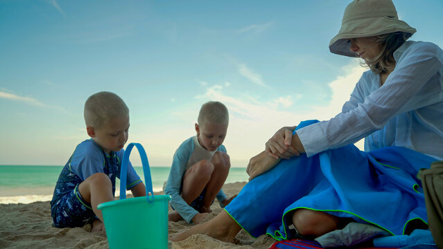 A mother watches her two young boys play in the sand at the beach. The sun is shining brightly, creating a cheerful atmosphere as they build sandcastles.