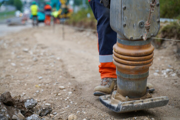Close-up of workers' legs using a vibrating construction plate to compress and level gravel,...