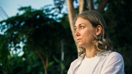 A woman stands amidst greenery, gazing thoughtfully into the distance. The warm light of the setting sun highlights her features while she reflects quietly in a tranquil environment.