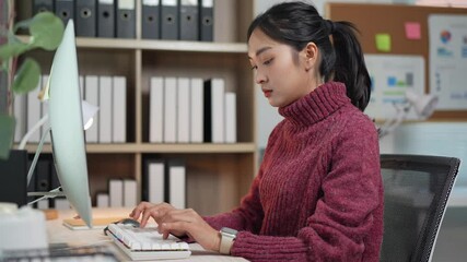 Young asian businesswoman wearing turtleneck sweater and smartwatch, focused on working, typing on a white keyboard in a modern office with organized shelves and charts on wall - Powered by Adobe