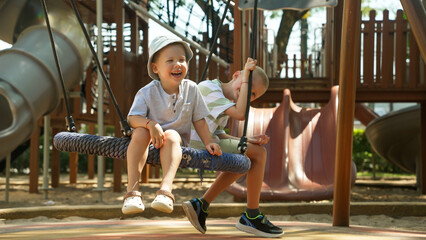 Two young children play at a playground, laughing and enjoying themselves on swings. One child swings joyfully while the other holds on tightly, surrounded by inviting play structures.