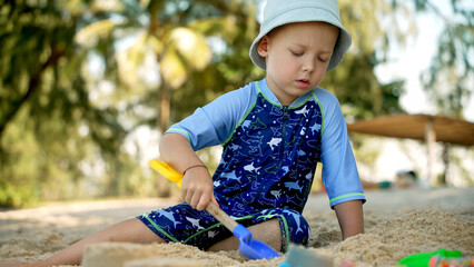 A young child wearing a blue sun hat and swimwear is focused on digging in the sand on a beach. The bright sunlight and palm trees create a cheerful and warm atmosphere.