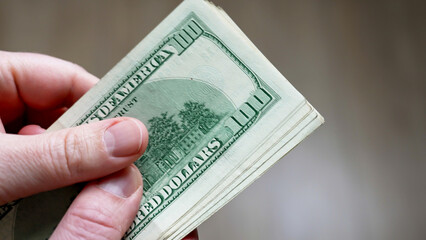 A person holds a bundle of crisp hundred dollar bills, showcasing the vibrant green hues. The hand is positioned in the sunlight, creating a clear focus on the money.