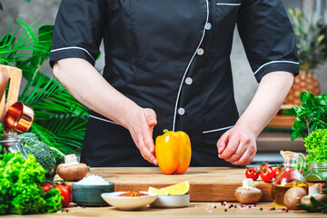 Chef in uniform holding orange paprika in hands.. Cozy kitchen with wooden table, kitchenware, vegetables, herbs and ingredients for cooking. Healthy vegan eating, culinary, recipes, food blogging 