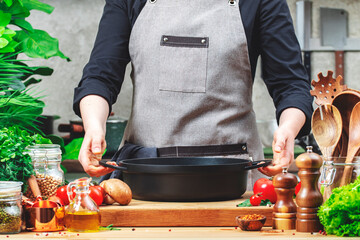 Housewife holds cast iron saucepan in hands. Cozy kitchen with wooden table, kitchenware,...