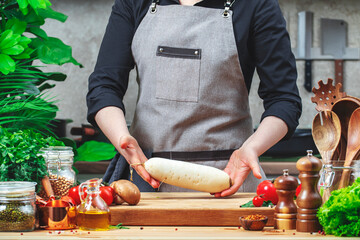 Cook holds organic white radish. Cozy kitchen with wooden table, kitchenware, vegetables, herbs and ingredients for cooking. Healthy vegan eating, culinary, recipes, blogging