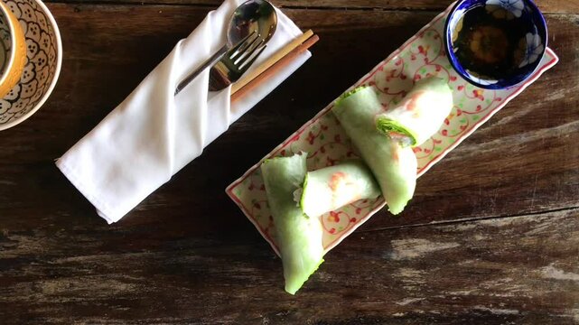 Fresh Vietnamese spring rolls, known as Gỏi cuốn, are served with shrimps and dipping sauce on a decorated plate, accompanied by cutlery and a napkin on a rustic wooden table in Hoi An, Vietnam