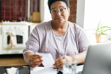 Upset black senior female in glasses looking at camera with sad facial expression holding paycheck or utility bill in hands sitting at kitchen table, worried of high prices for electricity