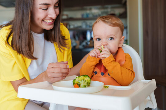 Happy mother with son eating broccoli sitting on high chair at home