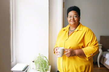Portrait of overweight elderly female of african ethnicity drinking healthy vitamin tea being on diet standing near window in bedroom, looking at camera with smile. Healthcare and wellness