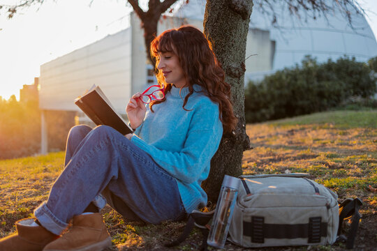 Smiling woman reading book and sitting near tree in park