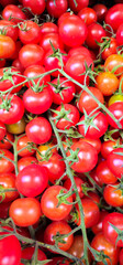 Red cherry tomatoes on green stems in a dense pile; some tomatoes have an orange hue, and the green sepals contrast with the smooth, shiny skin of the fruit;