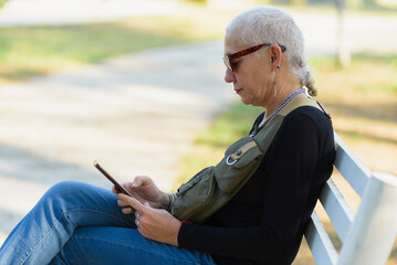 Fototapeta premium Middle-aged lesbian woman sitting on a park bench checks her cell phone.