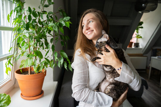 Smiling woman standing with Cornish Rex cat near potted plant on window