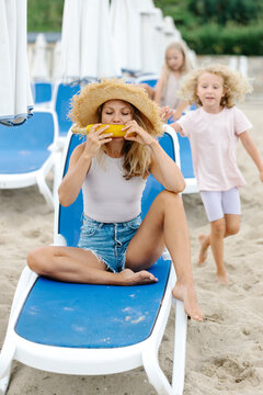 Woman sitting on lounger and eating corn cob near children at beach