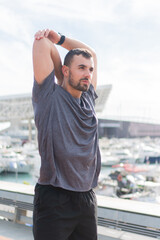 Focused Young Man Engaged in Dynamic Stretching Routine While Wearing Comfortable Sportswear at Outdoor Marina Setting with Boats in the Background for Fitness and Health Enthusiasts