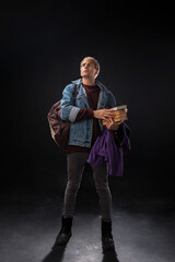 In a dimly lit studio, a person in a denim jacket and burgundy sweater raises a hand with intent. They hold books and a purple jacket, embodying youthful exploration.