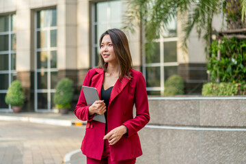 Fototapeta premium Confident Asian Businesswoman Holding Tablet Outdoors. Modern Career Woman in Red Suit in Urban Setting