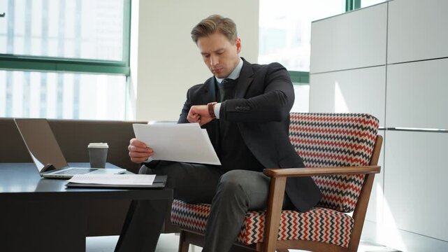 Stylish blond businessman in a dark suit checks time on his watch while reviewing documents sitting in a modern office space chair, laptop, and coffee cup on a sleek desk near large windows