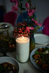 There is a candle, a vase of flowers and a plate of food on the table. The scene is warm and inviting, as the candlelight creates a cozy atmosphere