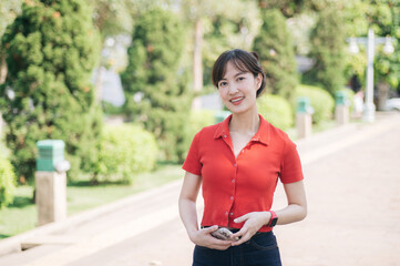 Asian cheerful woman in red shirt stands outdoors in park, holding phone, surrounded by greenery...