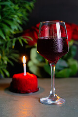 Red Cake with Candle and Wine Glass, Romantic Setting. Red cake with a lit candle, a glass of red wine, and blurred roses in the background. Ideal for romantic celebrations or anniversaries.