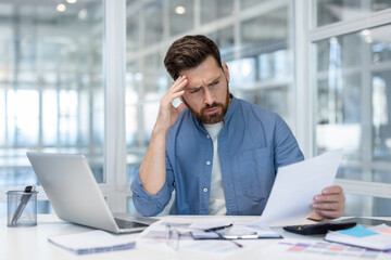 A concerned businessman reviews documents at his desk with a laptop in a bright, modern office...