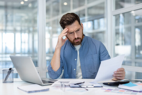 A concerned businessman reviews documents at his desk with a laptop in a bright, modern office setting, displaying stress about his finances
