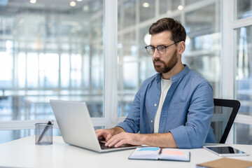 Fototapeta premium Serious thinking man working with laptop inside office at workplace, employee typing on keyboard.